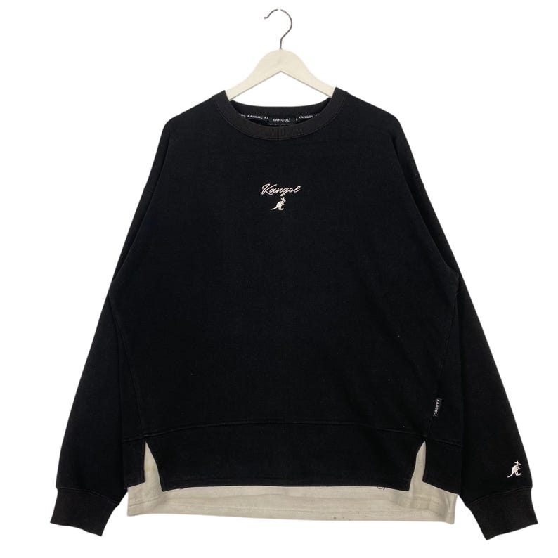 May include: Black sweatshirt with the white "Kangol" logo and kangaroo emblem. The sweatshirt has a layered design with a light beige underlayer. The long-sleeved top is hanging on a white hanger.