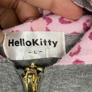 May include: Close-up of a gray hoodie with a gold zipper. The interior lining is pink with a pattern of dark pink hearts. A white label with the text "HelloKitty" is sewn into the garment.