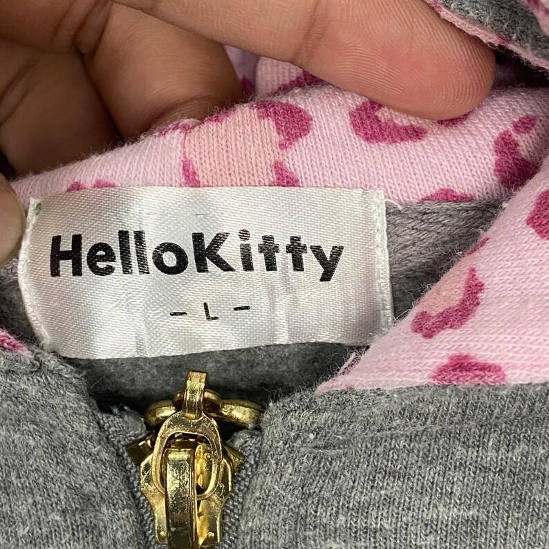 May include: Close-up of a gray hoodie with a gold zipper. The interior lining is pink with a pattern of dark pink hearts. A white label with the text "HelloKitty" is sewn into the garment.