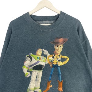 Op de afbeelding: Een donkerblauwe sweatshirt met een afbeelding van Buzz Lightyear en Woody uit de animatiefilm Toy Story. Buzz groet en Woody staat met zijn handen in zijn zij. De sweatshirt hangt aan een witte hanger.