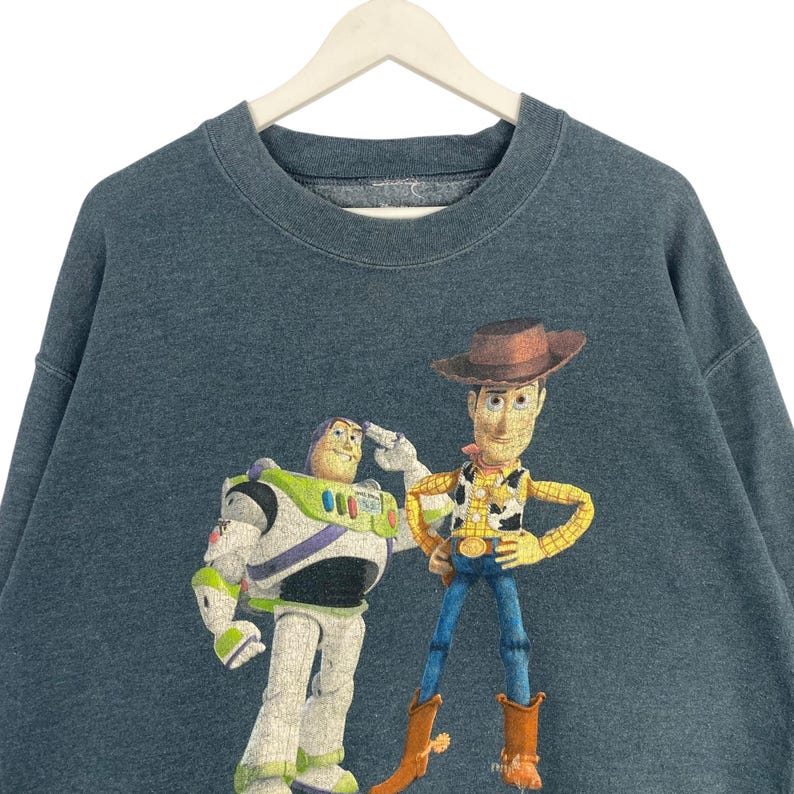 Op de afbeelding: Een donkerblauwe sweatshirt met een afbeelding van Buzz Lightyear en Woody uit de animatiefilm Toy Story. Buzz groet en Woody staat met zijn handen in zijn zij. De sweatshirt hangt aan een witte hanger.