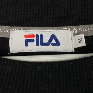 以下が含まれることがあります： 白いタグに青と赤の文字で「FILA」と書かれた黒いFilaのスウェットシャツ。タグには白いタグも付いており、そこに「M」の文字が書かれています。