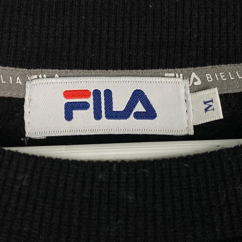 以下が含まれることがあります： 白いタグに青と赤の文字で「FILA」と書かれた黒いFilaのスウェットシャツ。タグには白いタグも付いており、そこに「M」の文字が書かれています。