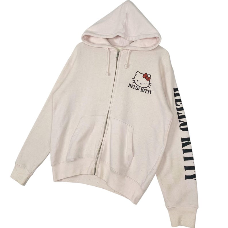 May include: Cream-colored zip-up hoodie featuring the Hello Kitty character with a red bow. The hoodie has a hood, a full-length zipper, and the words "HELLO KITTY" printed on the front and sleeve.