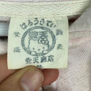 May include: Close-up of a light pink fabric tag with Japanese text and a circular graphic featuring a cartoon character. The tag is attached to a light pink garment.