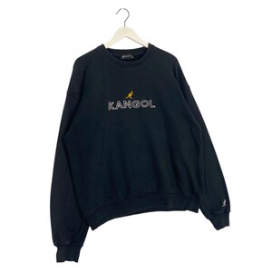 Puede incluir: Sudadera negra con la palabra "KANGOL" en blanco y un peque&ntilde;o logotipo de canguro amarillo encima. La sudadera tiene cuello redondo y mangas largas. Un peque&ntilde;o logotipo de canguro est&aacute; en la manga izquierda.