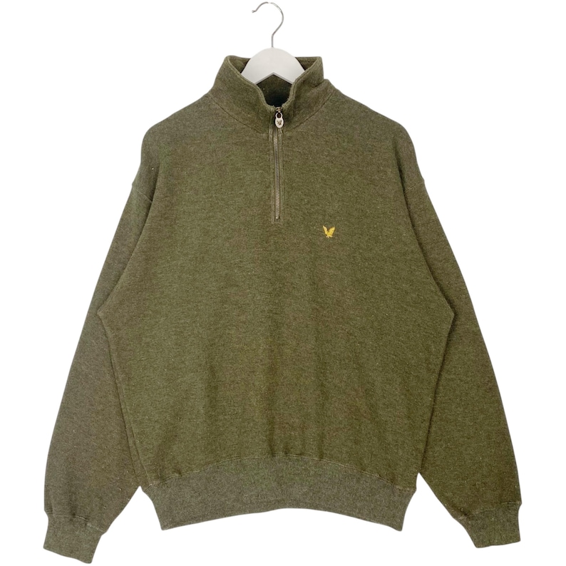 Puede incluir: Sudadera verde oliva con cierre de cremallera de cuarto de cuello y un peque&ntilde;o logotipo de p&aacute;jaro amarillo en el pecho. La sudadera tiene cuello alto y pu&ntilde;os y dobladillo acanalados. Colgada en una percha blanca.