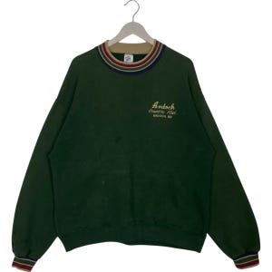 May include: A dark green sweatshirt with the text "Ardoch Country Club ARDOCH ND" embroidered in gold thread. The collar and cuffs have striped detailing in red, navy, and tan. The sweatshirt is hanging on a white hanger.