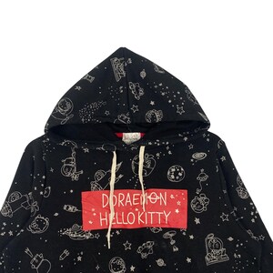 K&ouml;nnte beinhalten: Schwarzer Kapuzenpullover mit einem sich wiederholenden wei&szlig;en Muster aus Sternen, Planeten und Zeichentrickfiguren. Das Design umfasst Doraemon und Hello Kitty. Ein rotes Rechteck auf der Vorderseite zeigt den Text "DORAEMON HELLO KITTY". Der Hoodie hat wei&szlig;e Kordelz&uuml;ge.