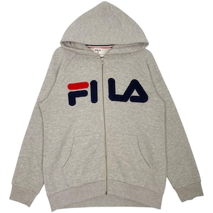 May include: A light gray zip-up hoodie with a hood and front pockets. The word "FILA" is prominently displayed on the front in a combination of red and navy blue. The hoodie has a full zipper and a small FILA logo on the inside of the hood.