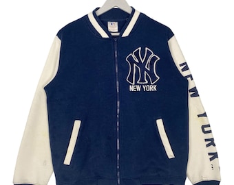 May include: A navy blue and white New York Yankees baseball jacket. The jacket features a full zip, white sleeves, and the team logo on the chest. The words "NEW YORK" are printed below the logo. The right sleeve has the words "NEW YORK" printed vertically.
