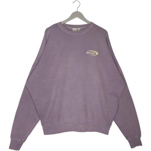 May include: A lavender-colored sweatshirt with the text "ROXY SURF" in yellow and white. The crew neck sweatshirt is hanging on a gray hanger. The long-sleeved top has a relaxed fit.