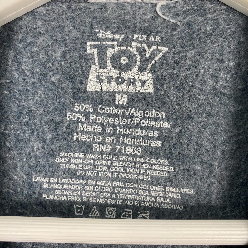 Op de afbeelding: Close-up van een grijs stoffen label met het witte Disney Pixar Toy Story logo. Het label bevat tekst met details over de stof samenstelling, herkomst en wasvoorschriften. Het label is bevestigd aan een grijs kledingstuk.
