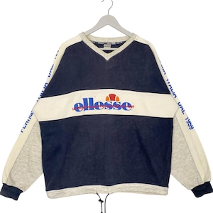 May include: A vintage navy blue and cream-colored sweatshirt with the brand name "ellesse" across the chest. The long sleeves are gray with the text "ITALIA DAL 1959" printed on the sleeves. The sweatshirt has a V-neck and a drawstring at the bottom.