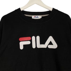 以下が含まれることがあります： 「F」の上部に赤いラインが入った白いFILAロゴがついた黒いスウェットシャツ。