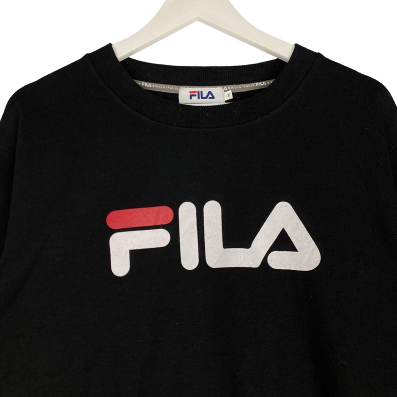 以下が含まれることがあります： 「F」の上部に赤いラインが入った白いFILAロゴがついた黒いスウェットシャツ。