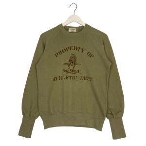 May include: Olive green sweatshirt with the text "PROPERTY OF" above an illustration of a seated figure, and "Paul Stuart ATHLETIC DEPT." below. The long-sleeved top has a crew neck and ribbed cuffs.