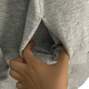 May include: A close-up of a gray sweatshirt with a pocket. The pocket is open and a hand is reaching into it.