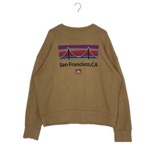 May include: A tan sweatshirt with a graphic of the Golden Gate Bridge in red, purple, and grey. The text "San Francisco. CA" is below the bridge image, with a small logo beneath it. The long-sleeved top is made of a soft material.
