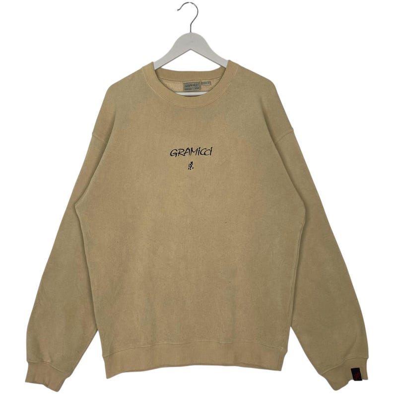 May include: A tan Gramicci sweatshirt with a crew neck and long sleeves, hanging from a white hanger. The Gramicci logo is printed in black on the chest. The sweatshirt has a faded appearance.