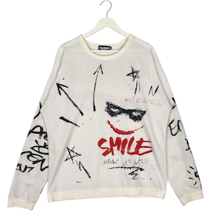 May include: White long-sleeve sweatshirt with a graphic design featuring a red smile, the word "SMILE", and the phrase "WHY SO SERIOUS?". The design also includes black arrows and scribbled text. The sweatshirt is hanging on a white hanger.