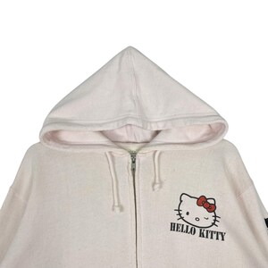 May include: A light pink zip-up hoodie featuring a hood and the Hello Kitty logo on the front. The logo includes a black outline of Hello Kitty's face, a red bow, and the words "HELLO KITTY" in black.