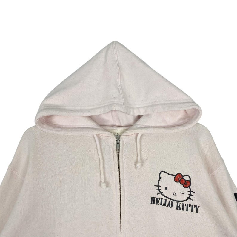 May include: A light pink zip-up hoodie featuring a hood and the Hello Kitty logo on the front. The logo includes a black outline of Hello Kitty's face, a red bow, and the words "HELLO KITTY" in black.