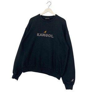 Puede incluir: Sudadera negra con la palabra "KANGOL" en letras blancas y un peque&ntilde;o logotipo de canguro dorado. La sudadera est&aacute; colgada de una percha blanca. Un peque&ntilde;o logotipo de canguro est&aacute; en la manga izquierda.