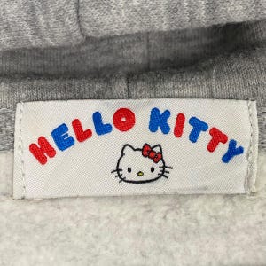 May include: A gray hooded sweatshirt with a white label that says "Hello Kitty" in red, blue, and white letters. A cartoon image of Hello Kitty is below the text.
