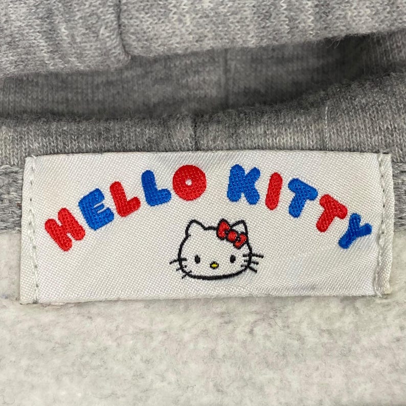 May include: A gray hooded sweatshirt with a white label that says "Hello Kitty" in red, blue, and white letters. A cartoon image of Hello Kitty is below the text.