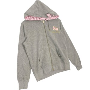 May include: A gray zip-up hoodie with a hood featuring pink leopard print lining and small ear details. The hoodie has a gold zipper and a small embroidered design with the text "Hello Kitty" in gold thread.