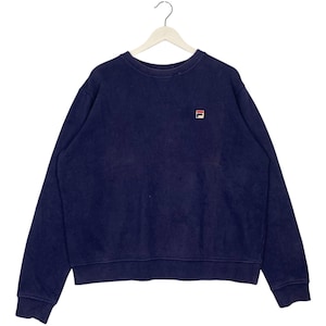 May include: Navy blue crew neck sweatshirt with a small logo on the upper left chest. The sweatshirt is hanging on a white hanger. The logo is a small square with red and white details. The sweatshirt has long sleeves and a relaxed fit.
