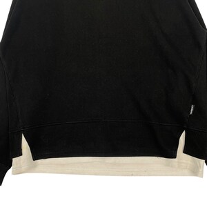 May include: Black sweatshirt with a white undershirt peeking out from the bottom. The sweatshirt has a crew neck and a split hem on the sides. The brand name "KANGOL" is visible on the right side of the sweatshirt.