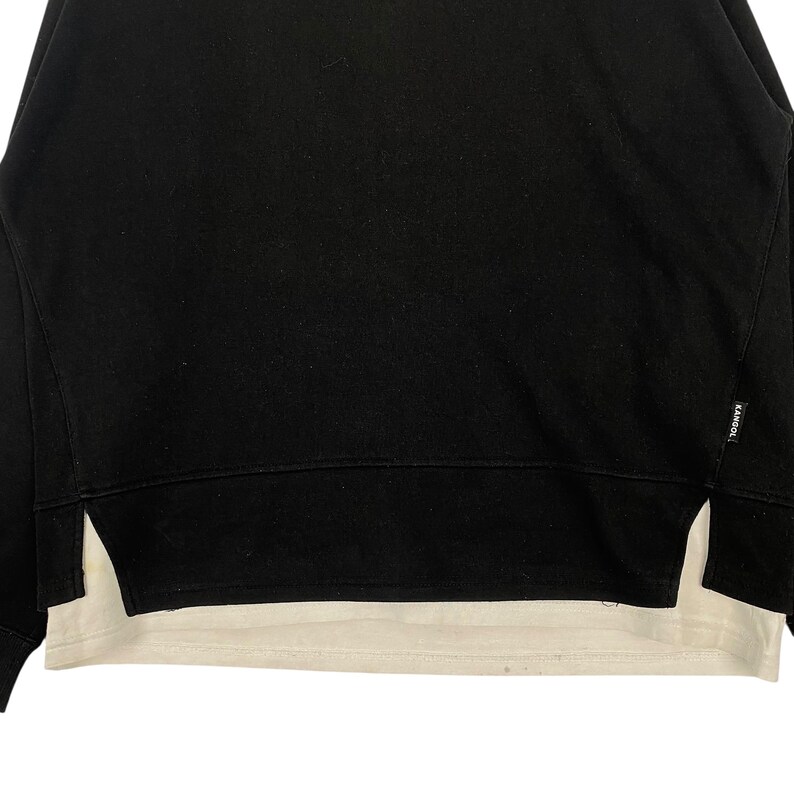 May include: Black sweatshirt with a white undershirt peeking out from the bottom. The sweatshirt has a crew neck and a split hem on the sides. The brand name "KANGOL" is visible on the right side of the sweatshirt.