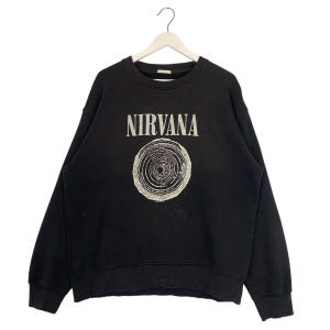 May include: Black crew neck sweatshirt with the word "NIRVANA" in white block letters above a circular graphic. The graphic is a detailed, off-white illustration of concentric circles and lines. The sweatshirt is hanging on a white hanger.