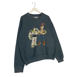 Op de afbeelding: Donker teal sweatshirt met een afbeelding van Buzz Lightyear en Woody uit Toy Story. Buzz draagt een wit en groen ruimtepak en groet. Woody draagt een cowboy outfit. De sweatshirt heeft een ronde hals en lange mouwen.