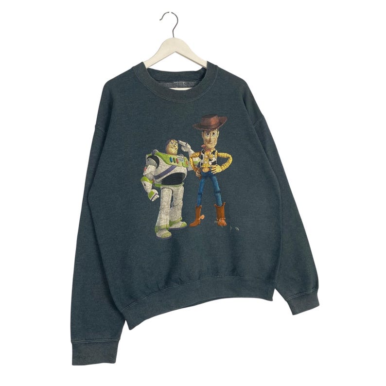 Op de afbeelding: Donker teal sweatshirt met een afbeelding van Buzz Lightyear en Woody uit Toy Story. Buzz draagt een wit en groen ruimtepak en groet. Woody draagt een cowboy outfit. De sweatshirt heeft een ronde hals en lange mouwen.