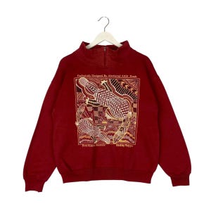 Puede incluir: Una sudadera roja con un gráfico de tres ornitorrincos en un patrón geométrico. El texto "Exclusively Designed By Aboriginal Artist Ponde" está por encima del gráfico. El texto "Perth Western Australia" y "Basking Platypus" está por debajo del gráfico.
