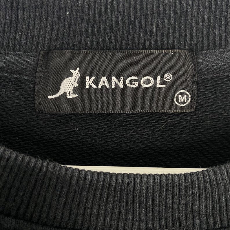 Puede incluir: Primer plano de un cuello de sudadera negro con una etiqueta con el nombre de la marca KANGOL en letras blancas. La etiqueta tambi&eacute;n muestra un logotipo de canguro blanco y un indicador de talla, la letra "M".