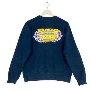 Puede incluir: Una sudadera azul marino de manga larga con un gráfico en la parte posterior. El diseño incluye el texto amarillo PIKO SPORTS sobre una flecha en espiral amarilla, rodeado por un borde de flores de color violeta claro y blanco.
