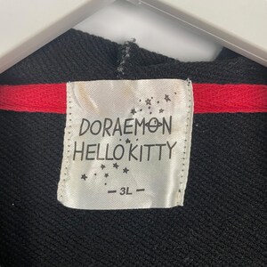 K&ouml;nnte beinhalten: Schwarzer Kapuzenpullover mit rotem Streifen und einem Etikett mit der Aufschrift "DORAEMON HELLO KITTY" mit der Gr&ouml;&szlig;e "- 3L -". Das Etikett hat Sterngrafiken.