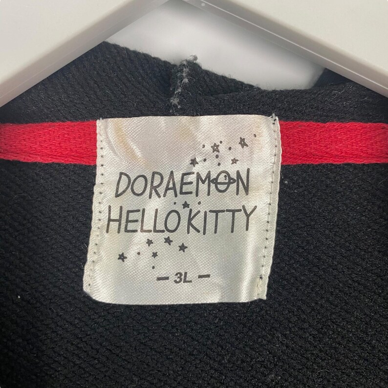 K&ouml;nnte beinhalten: Schwarzer Kapuzenpullover mit rotem Streifen und einem Etikett mit der Aufschrift "DORAEMON HELLO KITTY" mit der Gr&ouml;&szlig;e "- 3L -". Das Etikett hat Sterngrafiken.