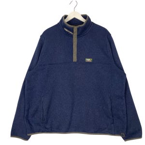 Könnte beinhalten: Ein marineblauer Fleece-Pullover mit hohem Kragen und Knopfleiste. Der Pullover hat eine kleine Tasche auf der linken Seite und einen kleinen Logoaufnäher auf der Brust. Der Kragen und die Blende sind mit einem kontrastierenden grauen Stoff versehen.