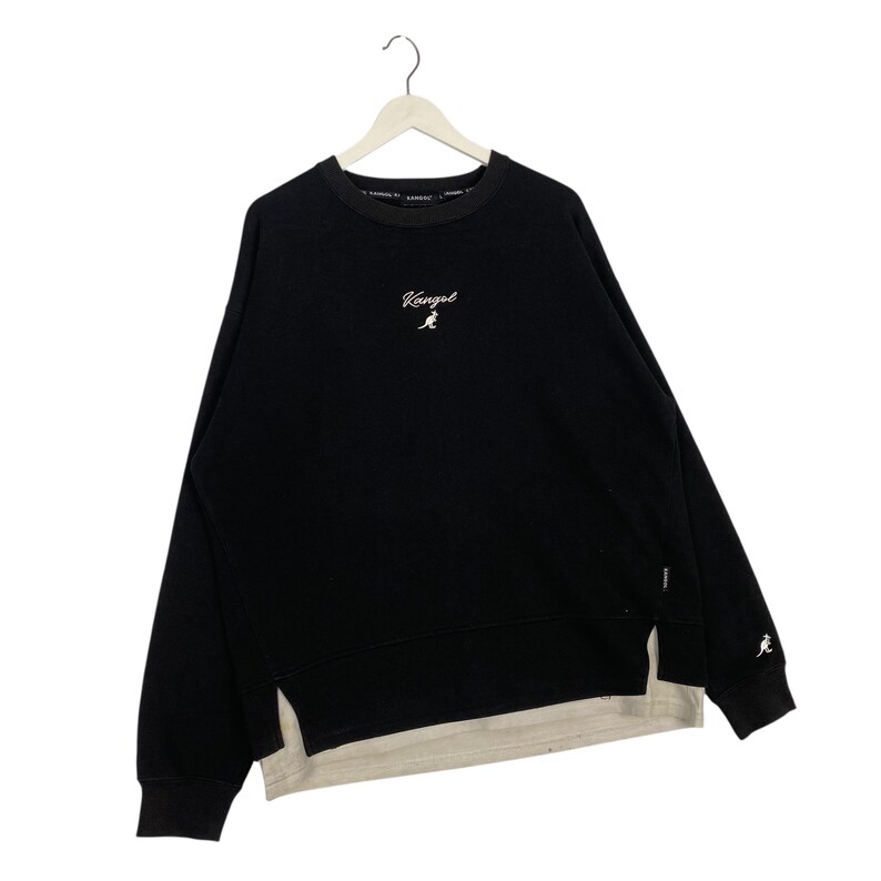 May include: Black sweatshirt with the word "Kangol" in white script on the chest. The sweatshirt has a layered design with a light beige underlayer. A small kangaroo logo is on the sleeve.