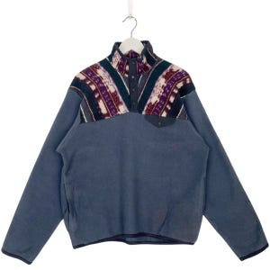 Könnte beinhalten: Ein blauer Fleece-Pullover mit einem gemusterten Joch mit lila, weißen und türkisfarbenen geometrischen Designs. Der Pullover hat einen Knopfkragen und eine kleine Tasche auf der Brust. Das langärmlige Oberteil hängt an einem weißen Kleiderbügel.