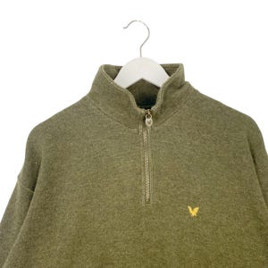 Puede incluir: Sudadera verde oliva con cremallera de cuarto de cuello y un peque&ntilde;o emblema dorado en el pecho. La sudadera tiene un cuello alto y una cremallera met&aacute;lica. La prenda est&aacute; colgada de una percha blanca.