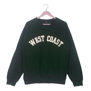 Puede incluir: Sudadera verde oscuro con la inscripción "WEST COAST" en blanco, arqueada sobre el pecho. La sudadera está colgada de una percha rosa. La prenda tiene mangas largas y cuello redondo.