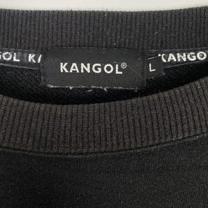 May include: Close-up of a dark gray sweatshirt collar with the brand name "KANGOL" in white text on a black tag. The tag also includes the registered trademark symbol. The sweatshirt has ribbed detailing.