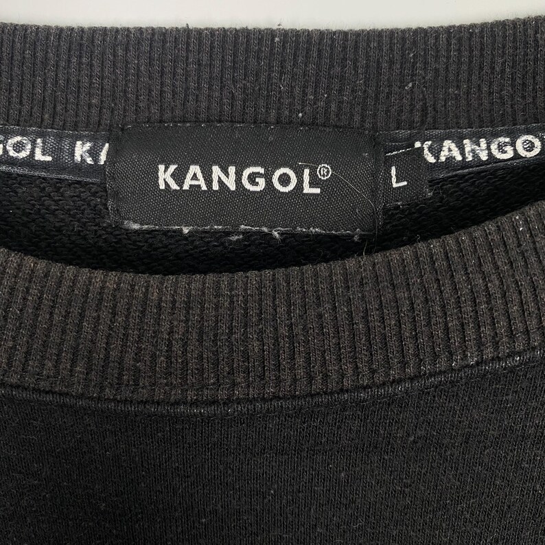 May include: Close-up of a dark gray sweatshirt collar with the brand name "KANGOL" in white text on a black tag. The tag also includes the registered trademark symbol. The sweatshirt has ribbed detailing.