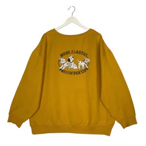 May include: Mustard yellow sweatshirt with a crew neck. The front features a graphic of four Dalmatian puppies with the text "MOST PLAYFUL FRIEND FOR LIFE" in an oval design. The sweatshirt is hanging on a white hanger.